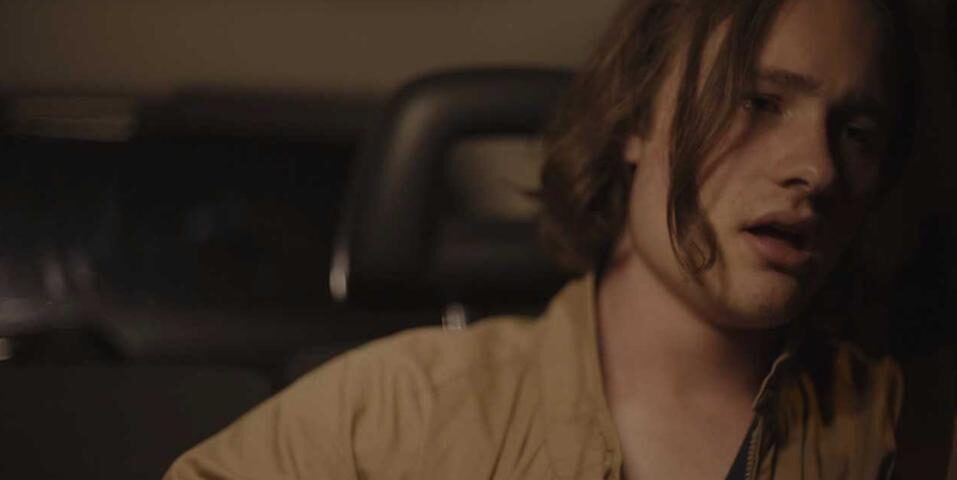 A young man sits alone in a car. He looks distraught.