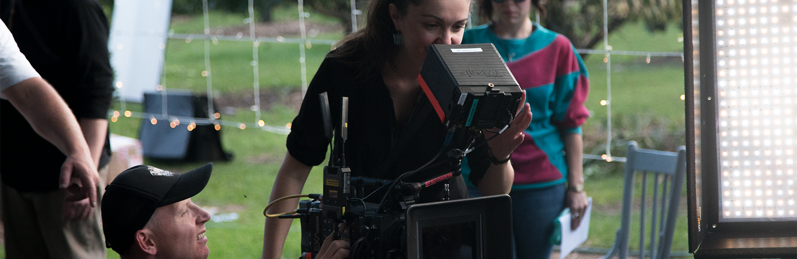 A cinematographer on location frames the scene through a camera's viewfinder as her camera assistant waits at the ready.