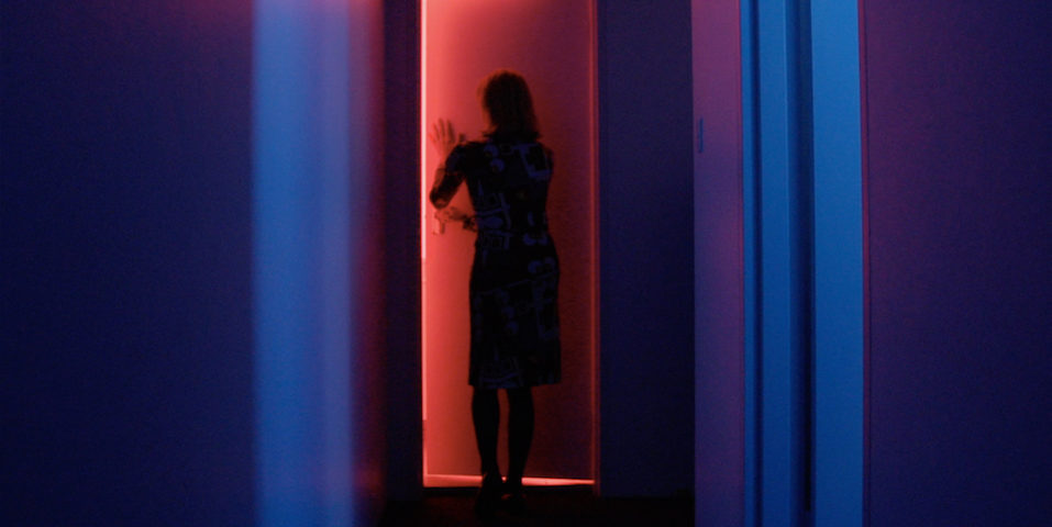 A woman in a room lit by blue lighting opens the door to a room lit with red lighting.