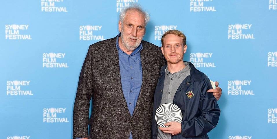Phillip Noyce and Tom Noaks take a photo together in front of the Sydney Film Festival photo backdrop. Noaks holds the Rouben Mamoulian Award for Best Director in his hands.