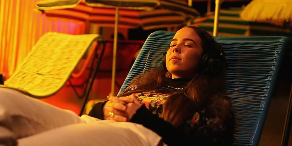 A young students sits peacefully with her eyes closed on a reclined folding beach chair with a set of earphones on in the Precipice art installation. The installation is dressed with beach umbrellas and more folding beach chairs and it's lit with orange lighting.