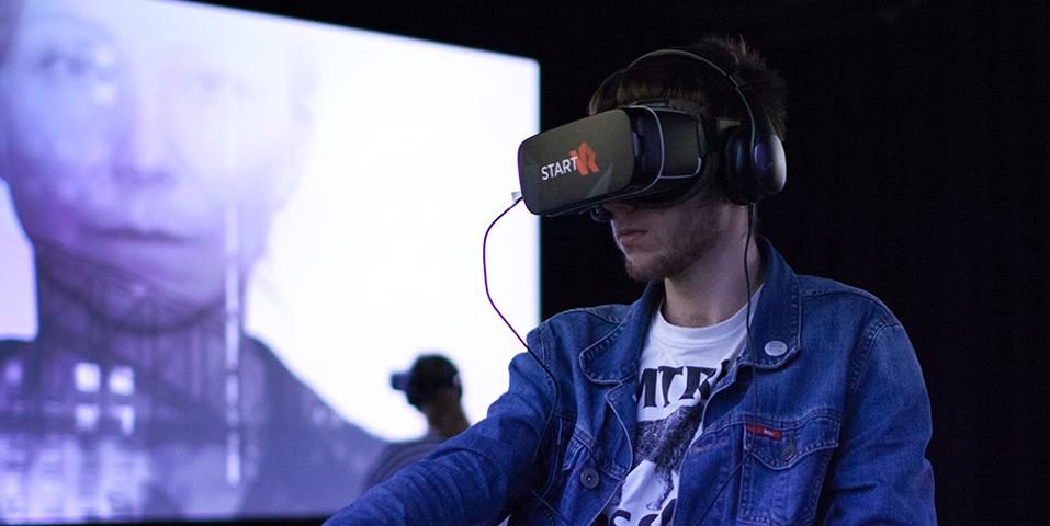 A young man sits in a studio with a Virtual Reality headset and headphones on. A design for the project's poster featuring the lead actress is projected onto a wall behind the man and the room is lit with indigo-coloured lighting.