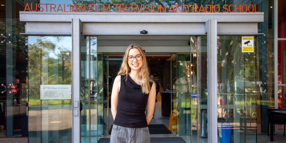 A person (Mia Channing) is standing in front of the entrance to the Australian Film Television and Radio School, smiling, with glass doors and the school's name visible above.